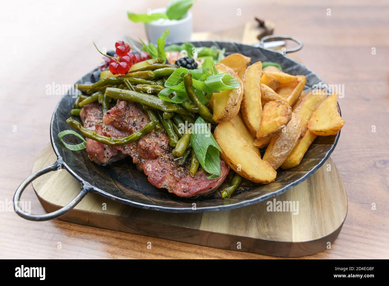 Rustic meal with roasted steak, potato wedges, green beans and a mixed salad with fruits, served in an iron cooking pan on a wooden table, selected fo Stock Photo
