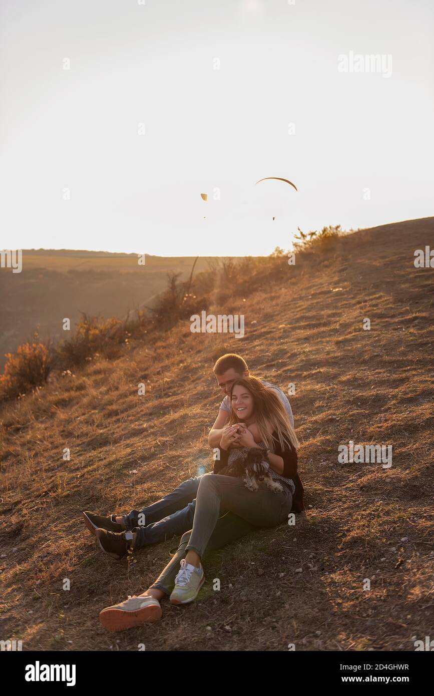 A young man hugs a beautiful girl from behind, the couple admire the sunset from the top of the hill. Happy family travels with a dog schnauzer. Stock Photo