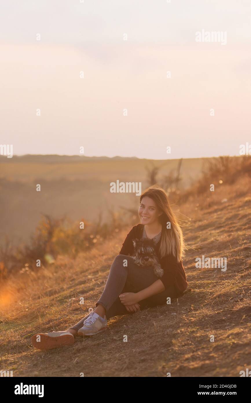 Happy young woman travels with her domestic dog black schnauzer. A beautiful girl hugs a pet, sits on a mountain, the wind develops her red long hair, Stock Photo