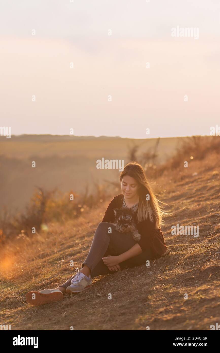 Happy young woman travels with her domestic dog black schnauzer. A beautiful girl hugs a pet, sits on a mountain, the wind develops her red long hair, Stock Photo