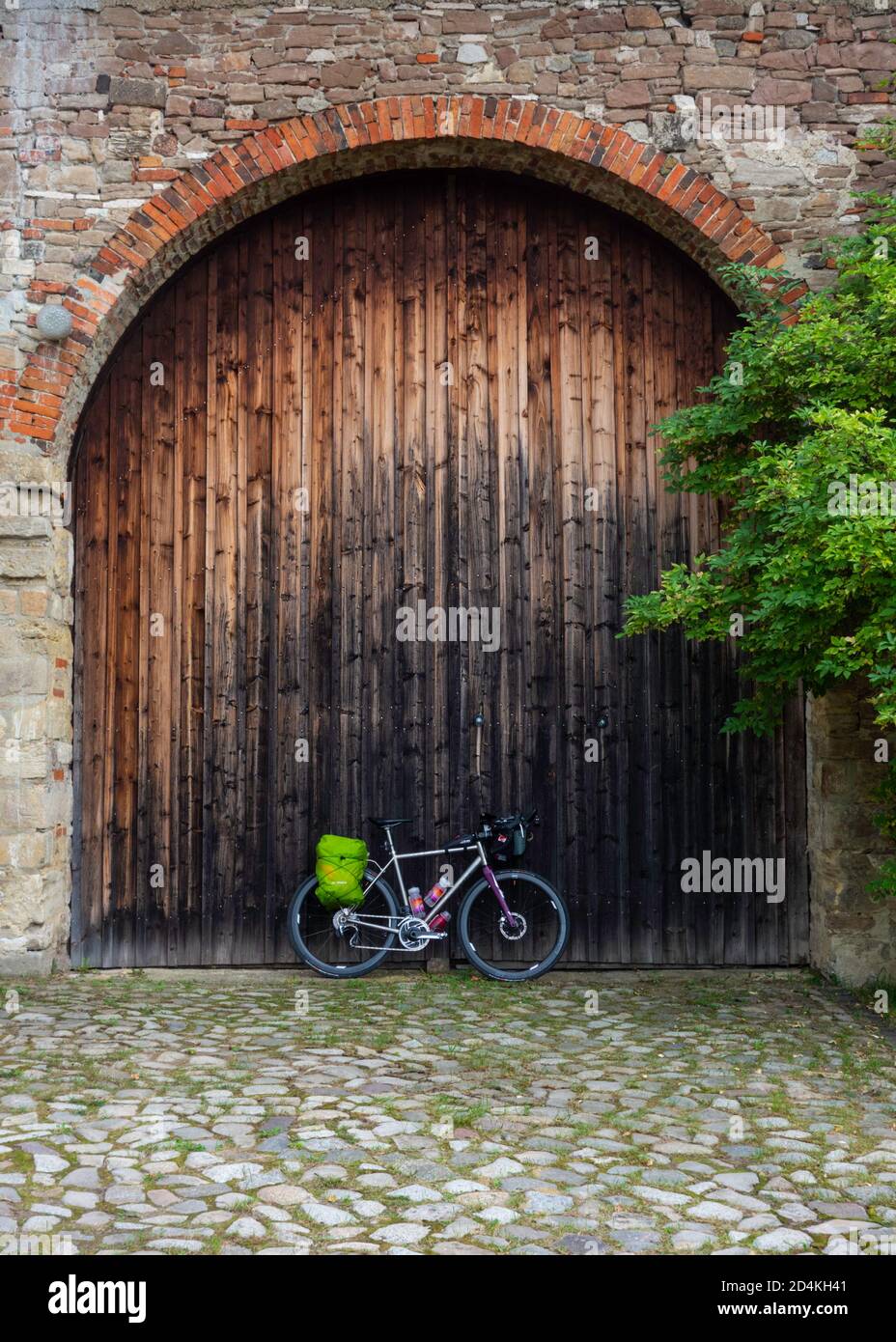 A titanium bike with drop bars and panniers parked in front of a big wooden gate surrounded by quarry stone masonry and cobbles. Thale, Harz region. Stock Photo