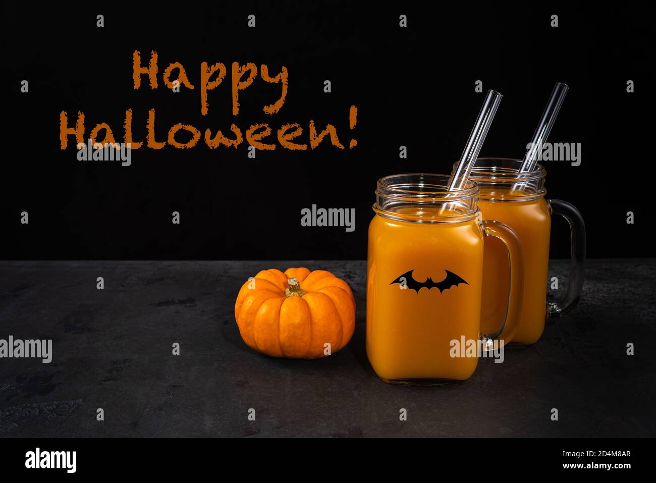 Two square glass jars with black bat. Non-alcoholic orange pumpkin mocktail and glass straws on black textured surface. Whole pumpkin is near jars. Ha Stock Photo