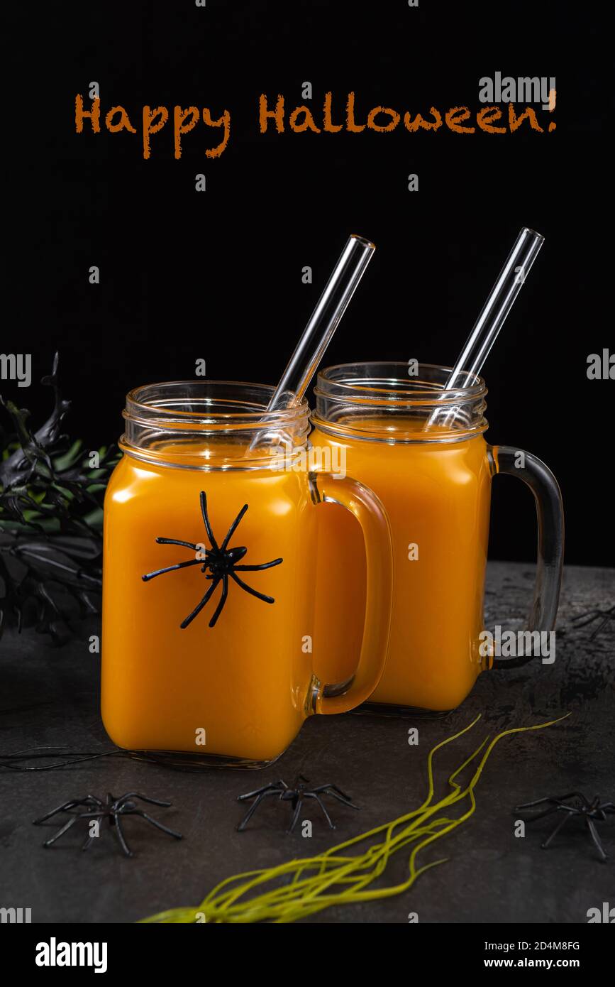 Two square glass jars with pumpkin mocktail and glass straws on black textured table decorated with spiders and spooky plants. Happy Halloween greetin Stock Photo