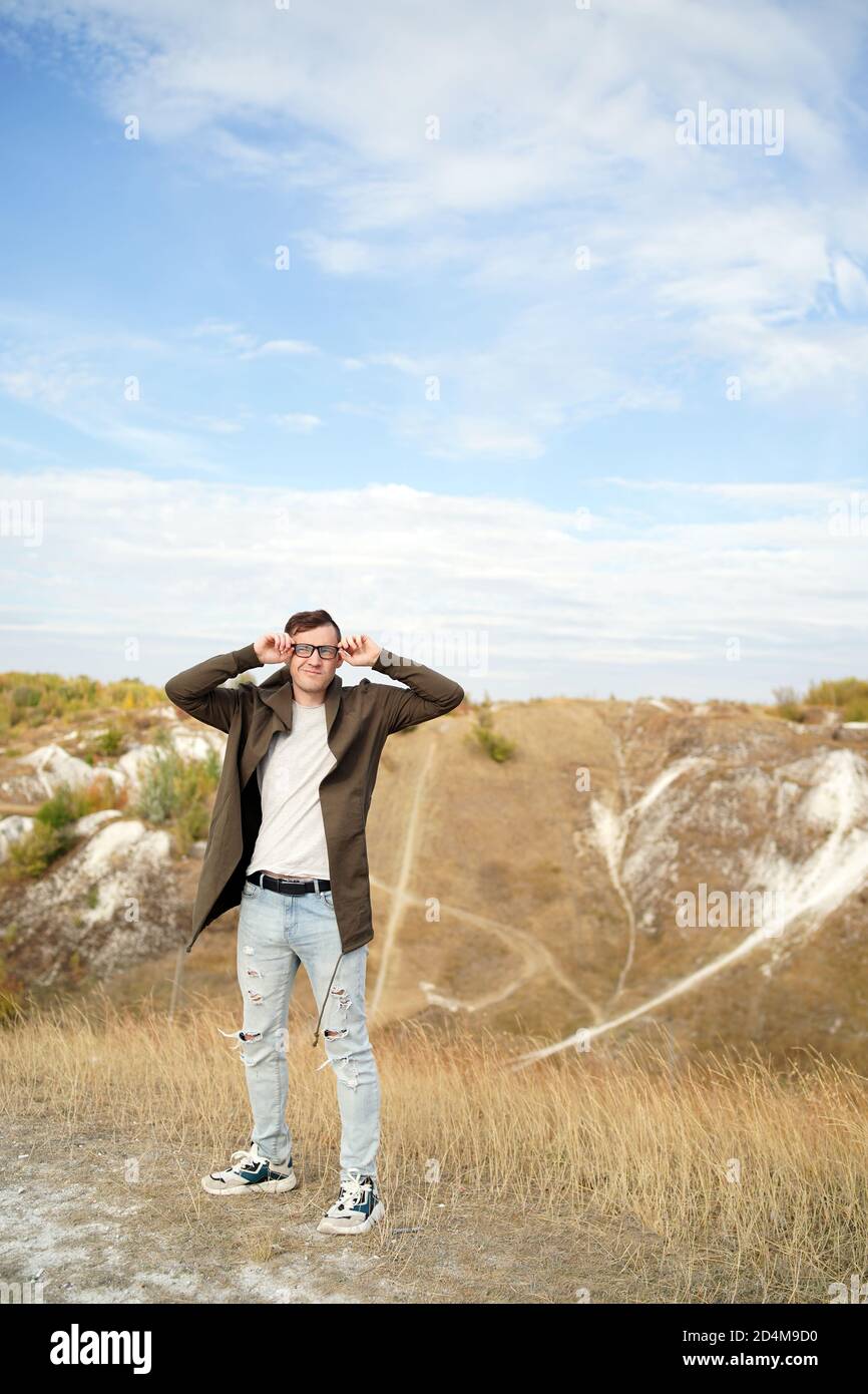 Man with glasses in jeans, t-shirt and cape stands in hilly terrain. Adult male in hood adjusts his eyeglasses and enjoys beautiful view of Stock Photo