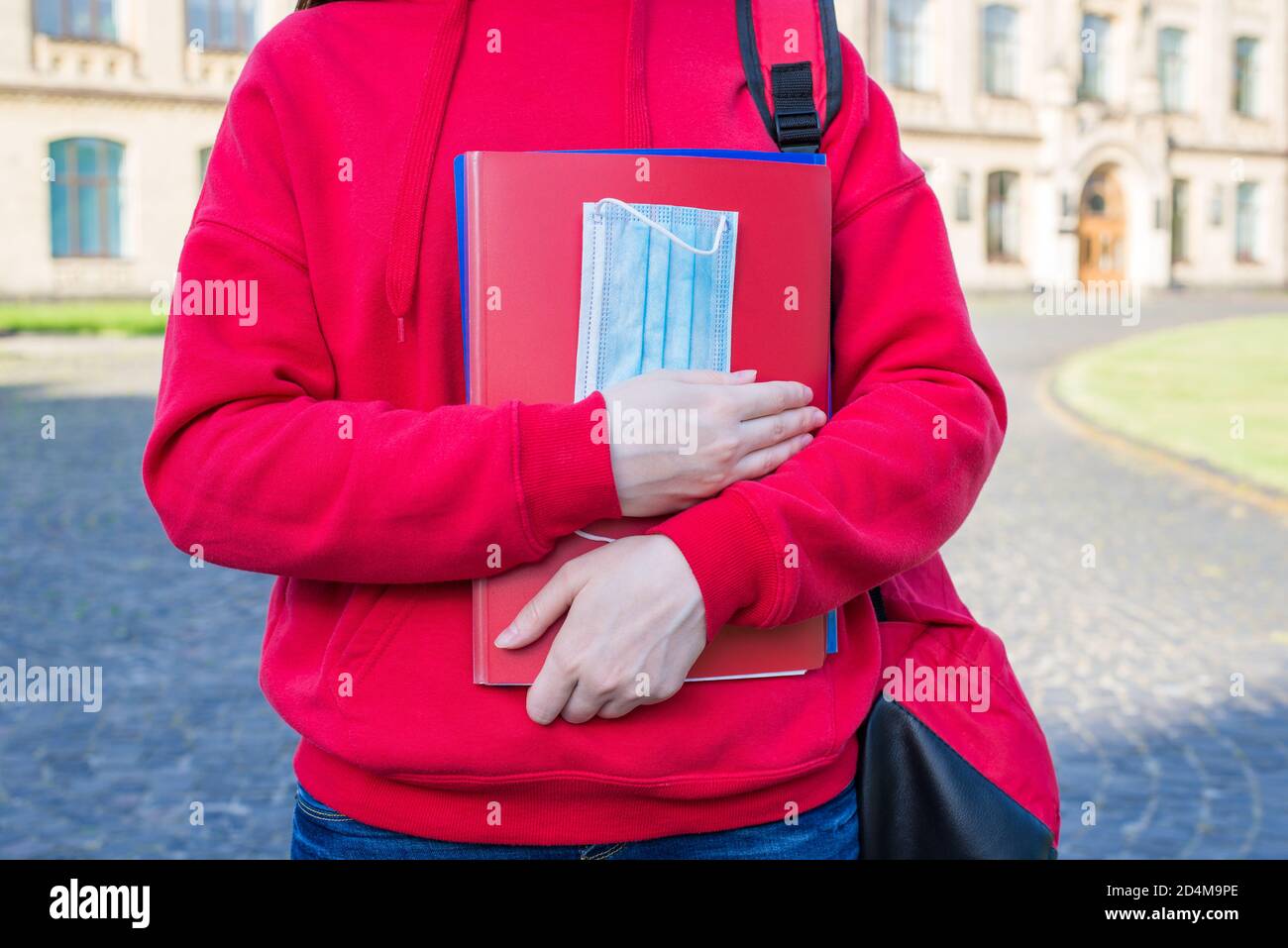 Cropped close up photo of smart clever teenager in casual red jumper holding notebooks and medical mask in hands Stock Photo