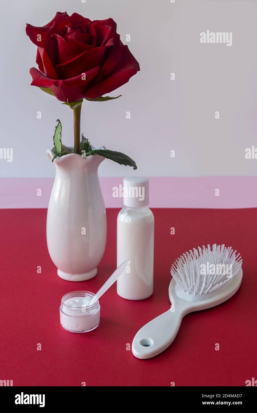 White hair comb, unlabeled shampoo bottle, hair care products and red rose in white vase on the table. Conception of natural cosmetics, spa treatments Stock Photo