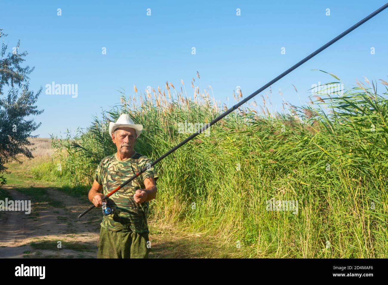 An old fisherman prepares fishing rod in summer on the river bank in reeds.  An elderly man in a white hat enjoys sport fishing. Bright sun, soft focus  Stock Photo - Alamy