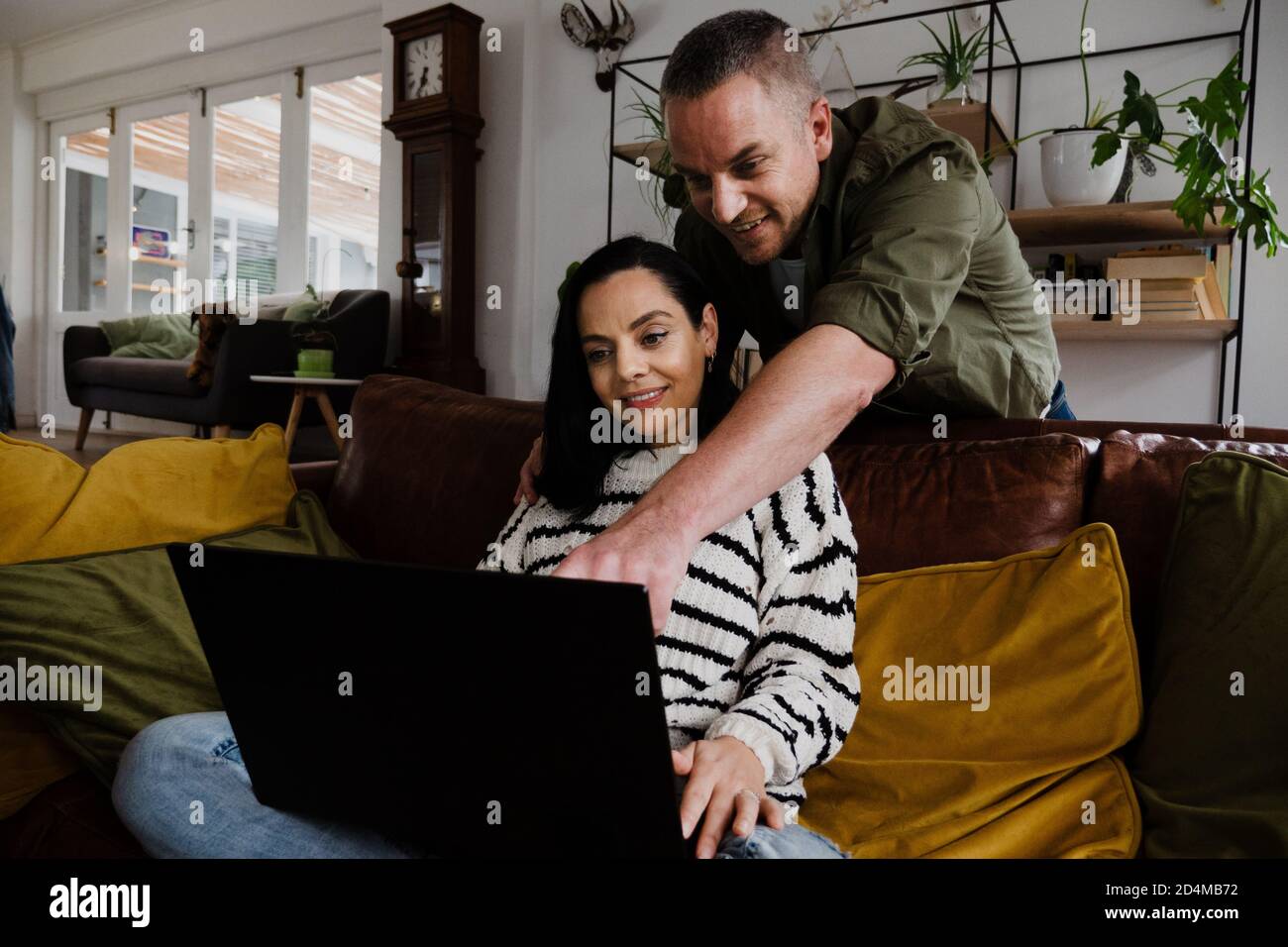 Handsome husband pointing at laptop while wife sits on comfy sofa in living room. Stock Photo