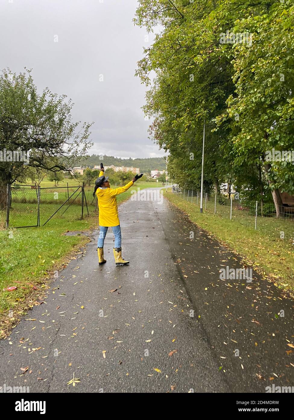 Happy woman in yellow raincoat and rain boots with jaguar pattern walking on rainy day. Back view of person. Stock Photo