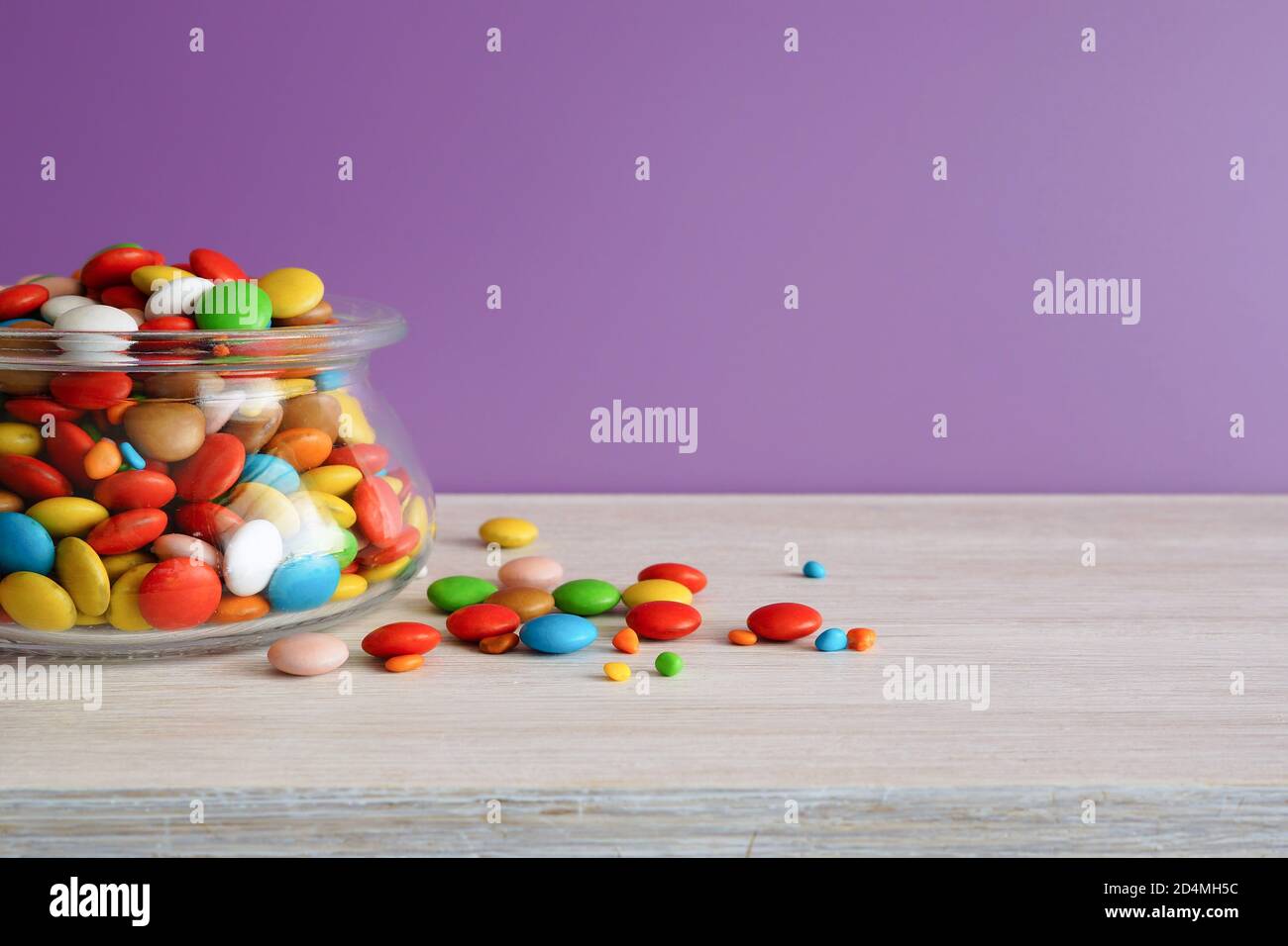 Multi-colored candies. Round candies in a glass jar on a white table. Lilac background Stock Photo