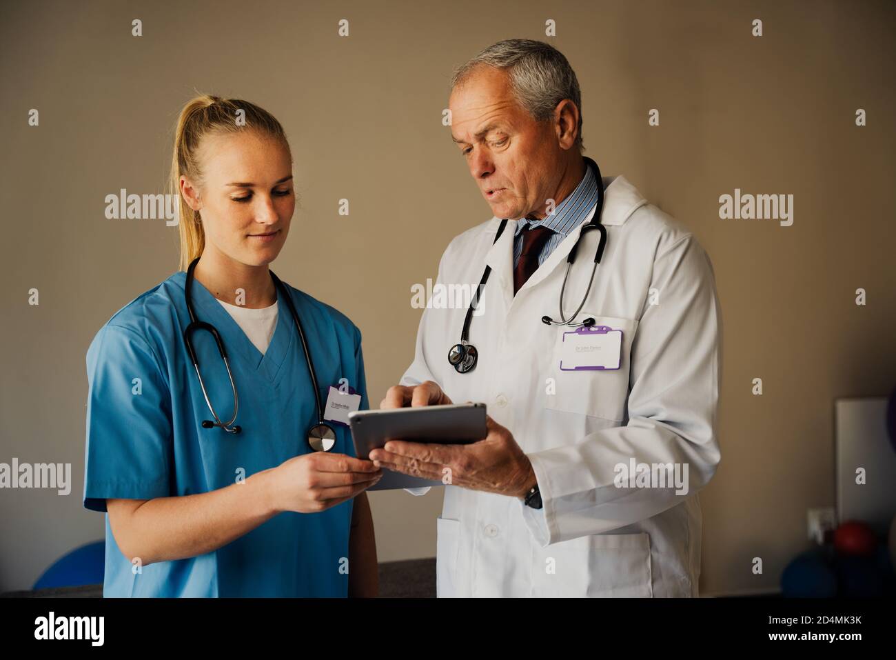 Male doctor teaching female intern in scrubs, while showing instructions on digital tablet in organised doctors room Stock Photo