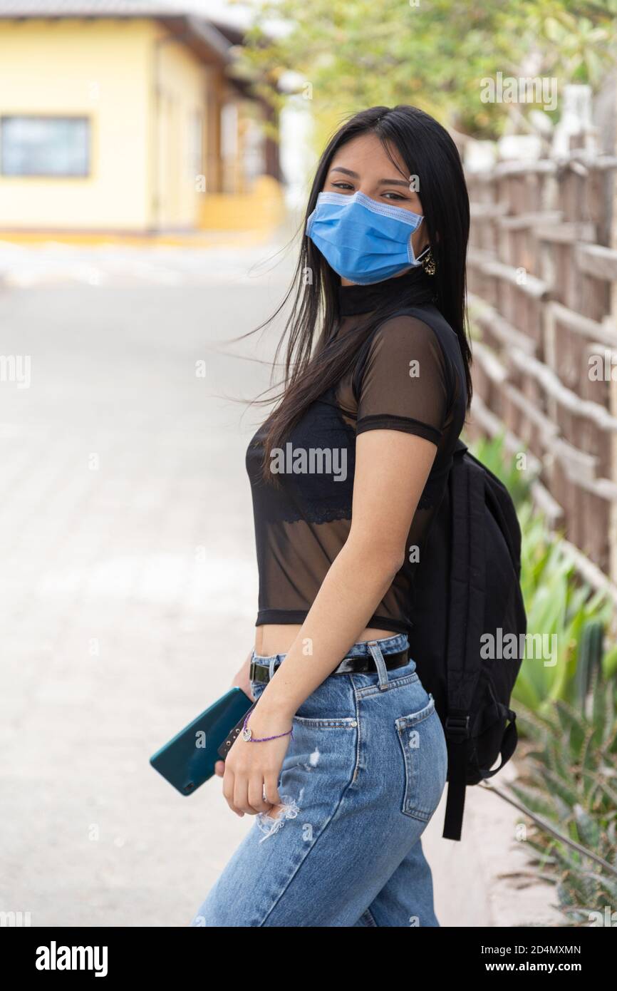 beautiful young latin woman with long black hair, sideways and with a cell phone in her hand, wearing a mask, transparent blouse with jeans Stock Photo