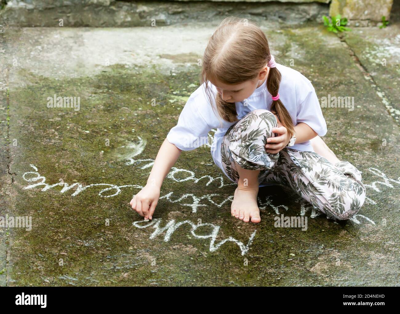 Small young girl writing simple words on concrete with white chalk. Child drawing with chalk on the ground outdoors. Art therapy concept. School kid Stock Photo
