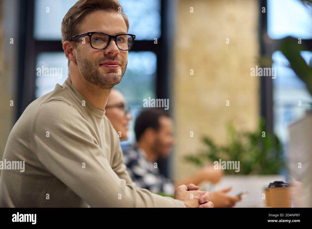 Young handsome man, male office worker sitting at desk in the board room and looking at camera while having a meeting with colleagues. Business people and office life, teamwork Stock Photo