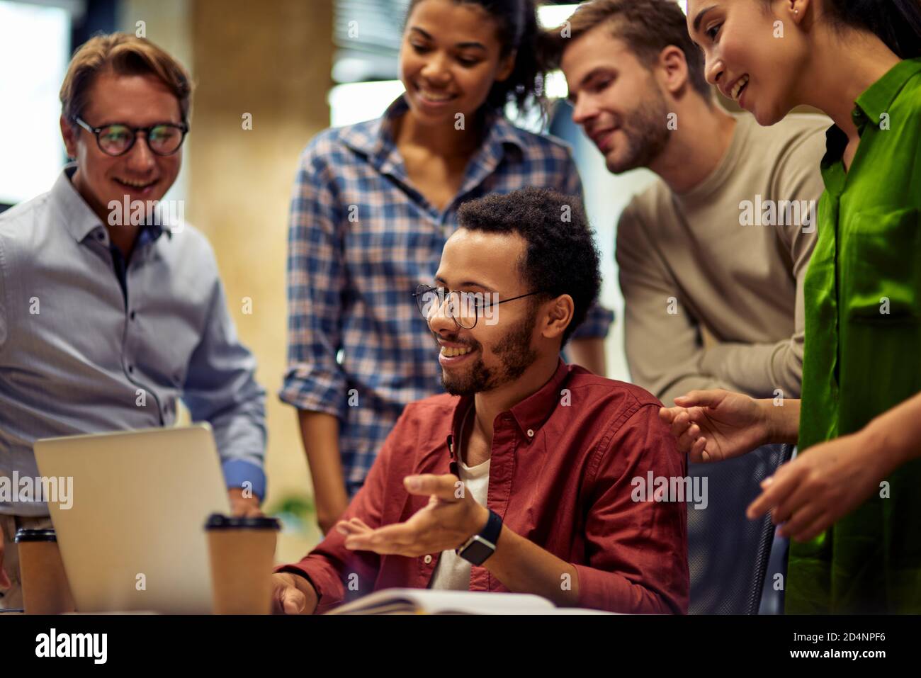 Working as a team. Group of happy multiracial business people looking at laptop screen and discussing work or project results while standing in the modern office or coworking space Stock Photo