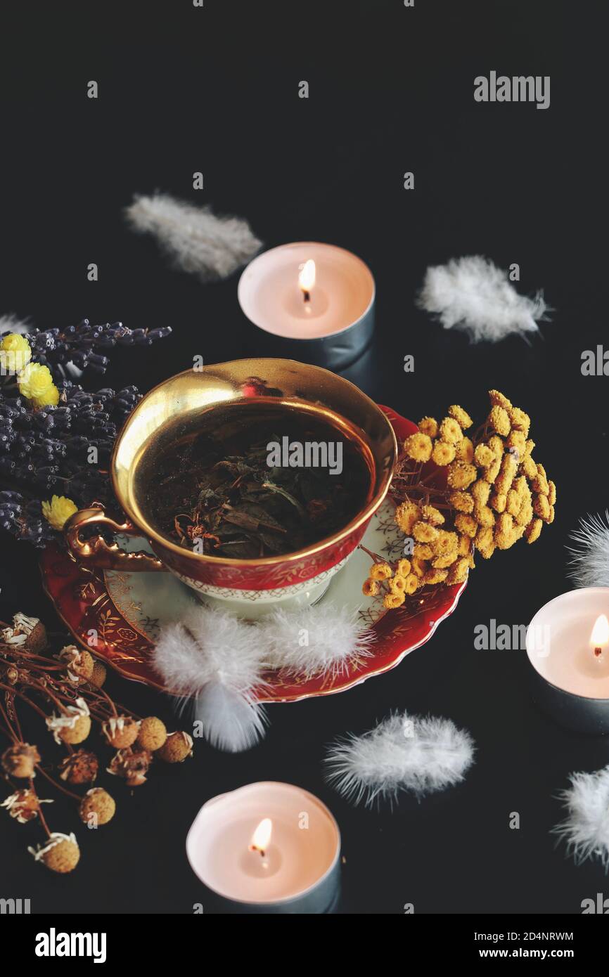 Vintage white red and gold teacup with green herbs in it on a dark black table. Dried chamomile, lavender, yellow flowers , burning lit candles Stock Photo