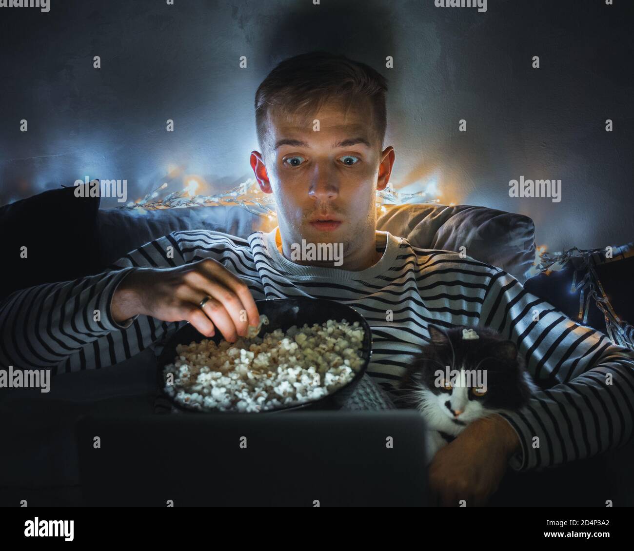 young man with black cat watching a movie eating popcorn on TV at home. Movie night. Relax,rest watching a horror film or video on screen. Background Stock Photo