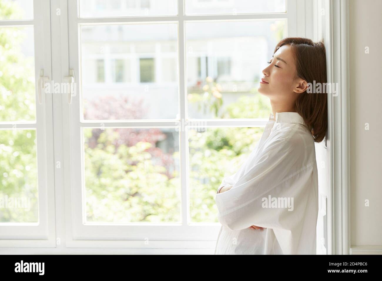 beautiful young asian woman standing by window at home arms crossed eyes closed Stock Photo