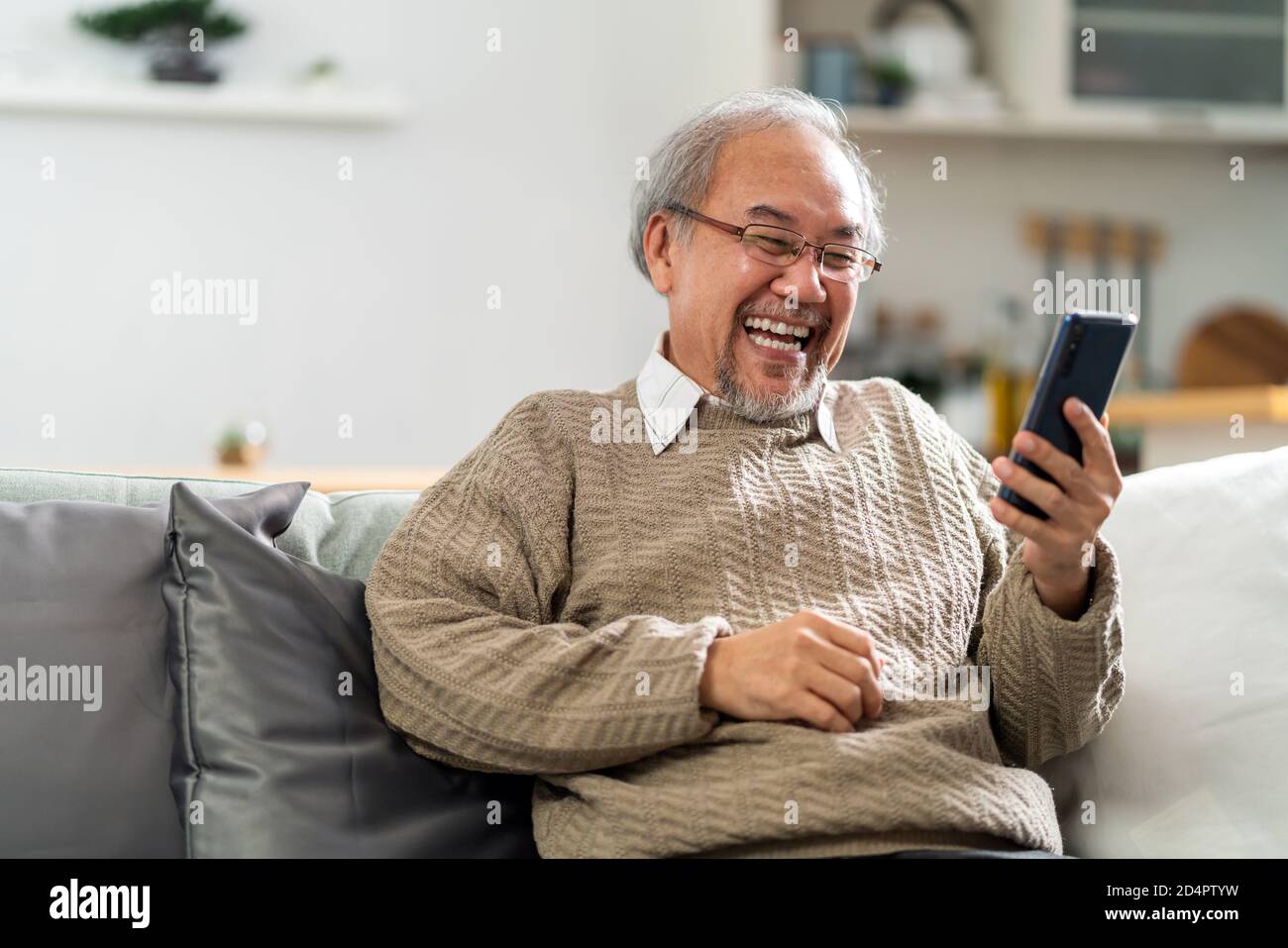 Happy retirement elderly man sitting on sofa at living room use cellphone to make video call to his family with laugh and smile. Communication Technol Stock Photo