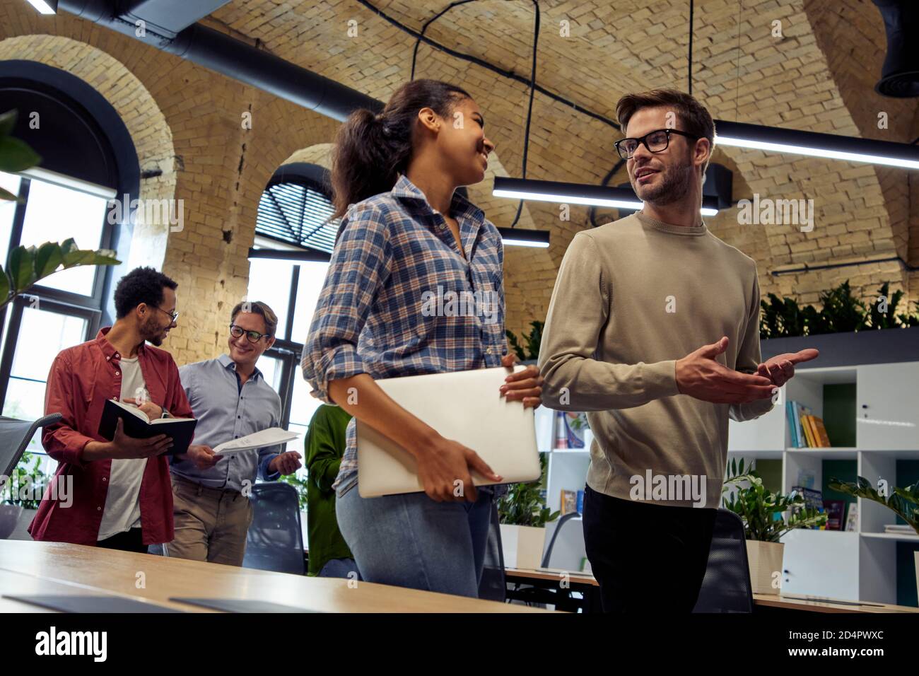 Informal conversation at work. Two young diverse business people, male and female coworkers talking about something while walking in the modern office. Business concept, teamwork and cooperation Stock Photo