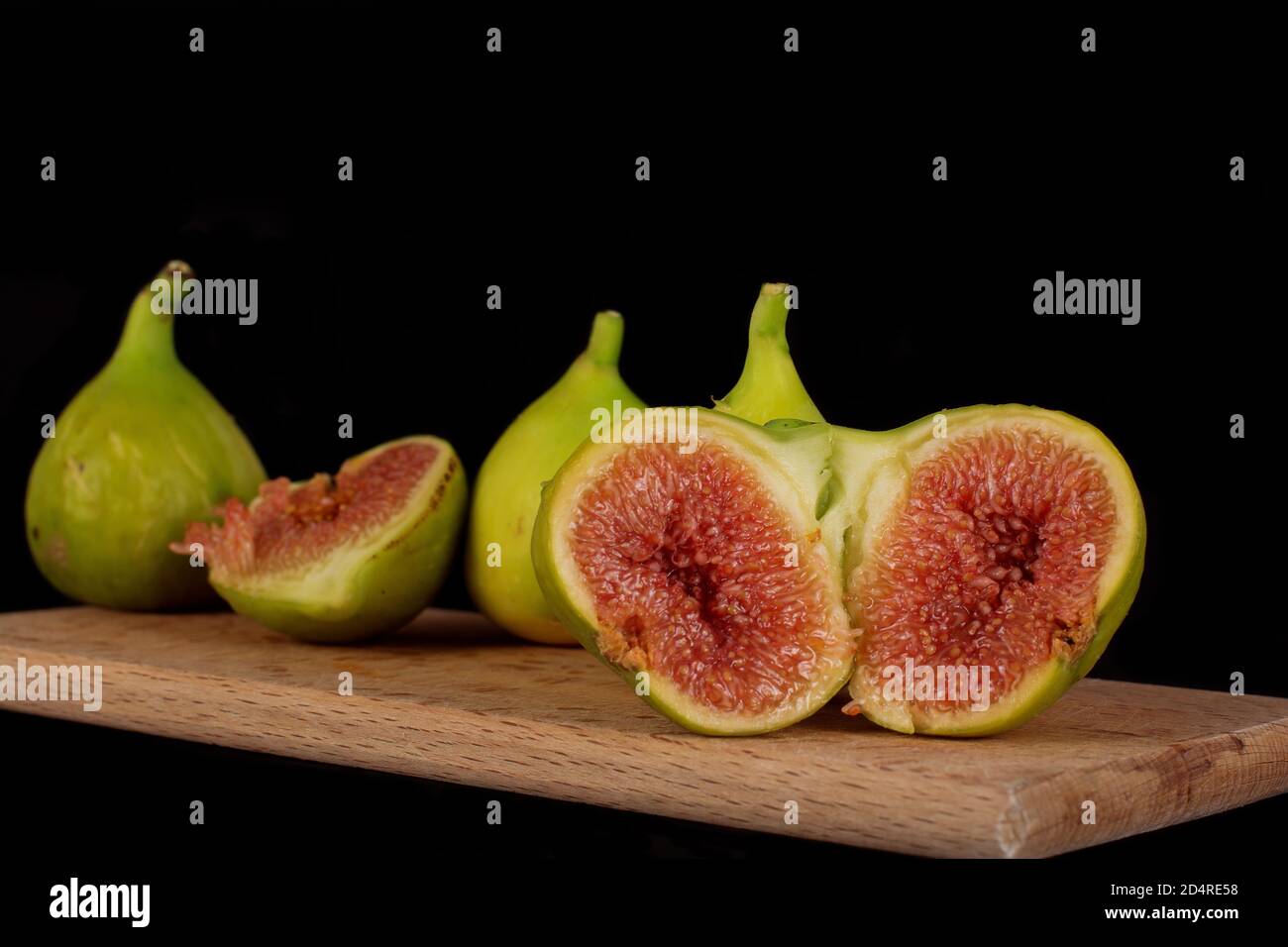Ripe fresh figs are lying on a wooden cutting board. Two halves of a common fig syconium are lying in the front plane, showing its one-seeded fruits. Stock Photo