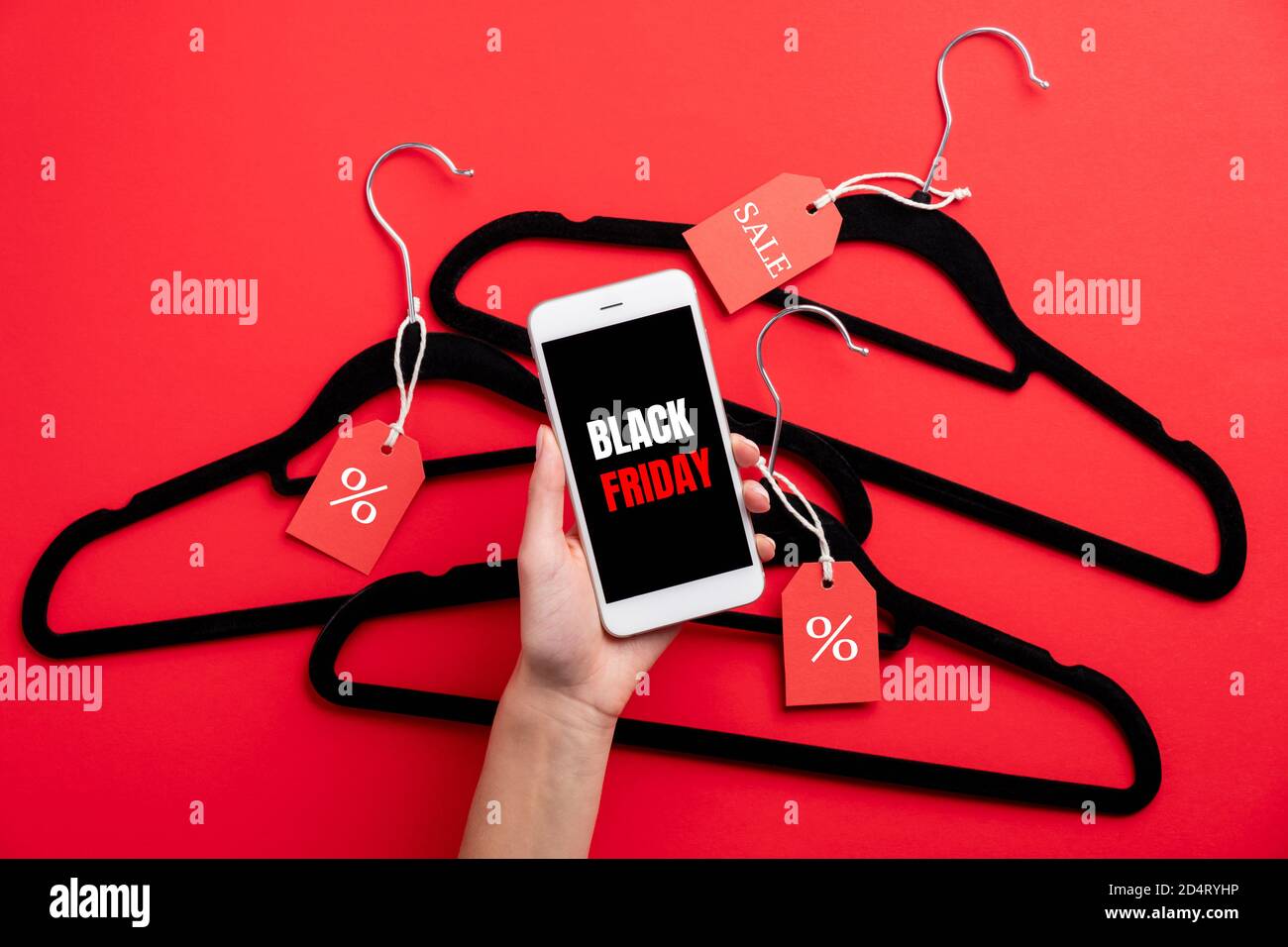Female hand holding phone with sign 'Black Friday' on screen over coat hangers on red background. Black Friday web shopping, coupon, discount concept. Stock Photo