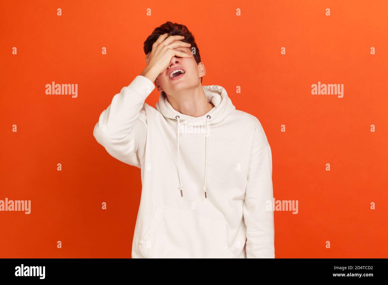 Depressed young man in casual style white hoodie closing eyes with arm, making facepalm gesture, loser, forget about something important. Indoor studi Stock Photo