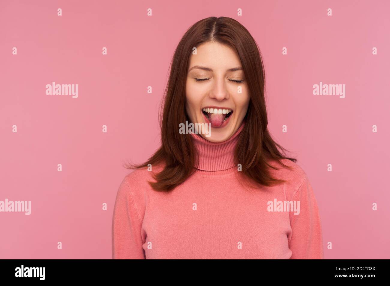 Closeup positive naughty woman with brown hair in pink sweater showing tongue standing with closed eyes, bragging, having fun, childish behaviour. Ind Stock Photo