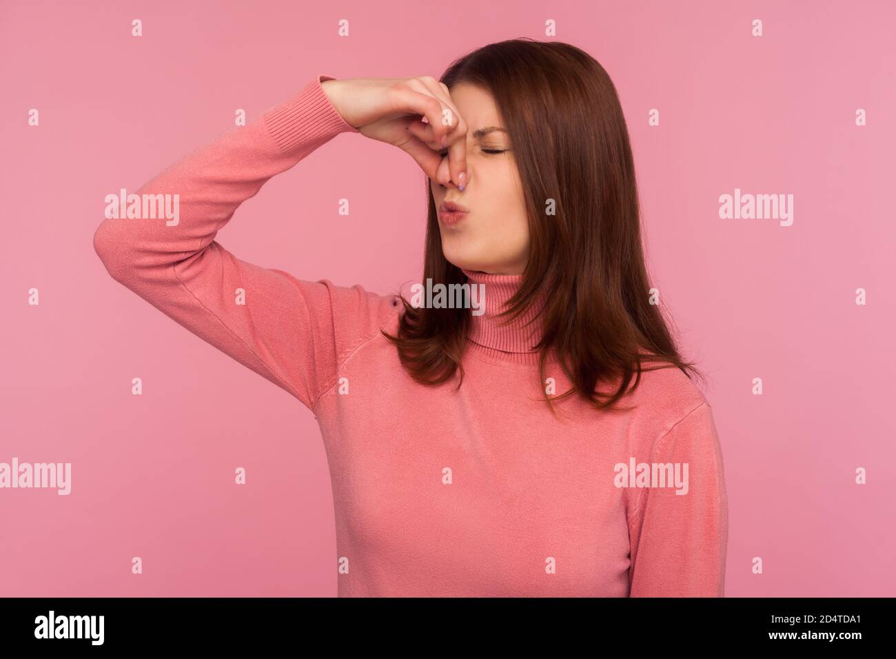 Unhappy confused brunette woman grabbing nose with fingers feeling unpleasant smell, shocked with disgusted fart. Indoor studio shot isolated on pink Stock Photo