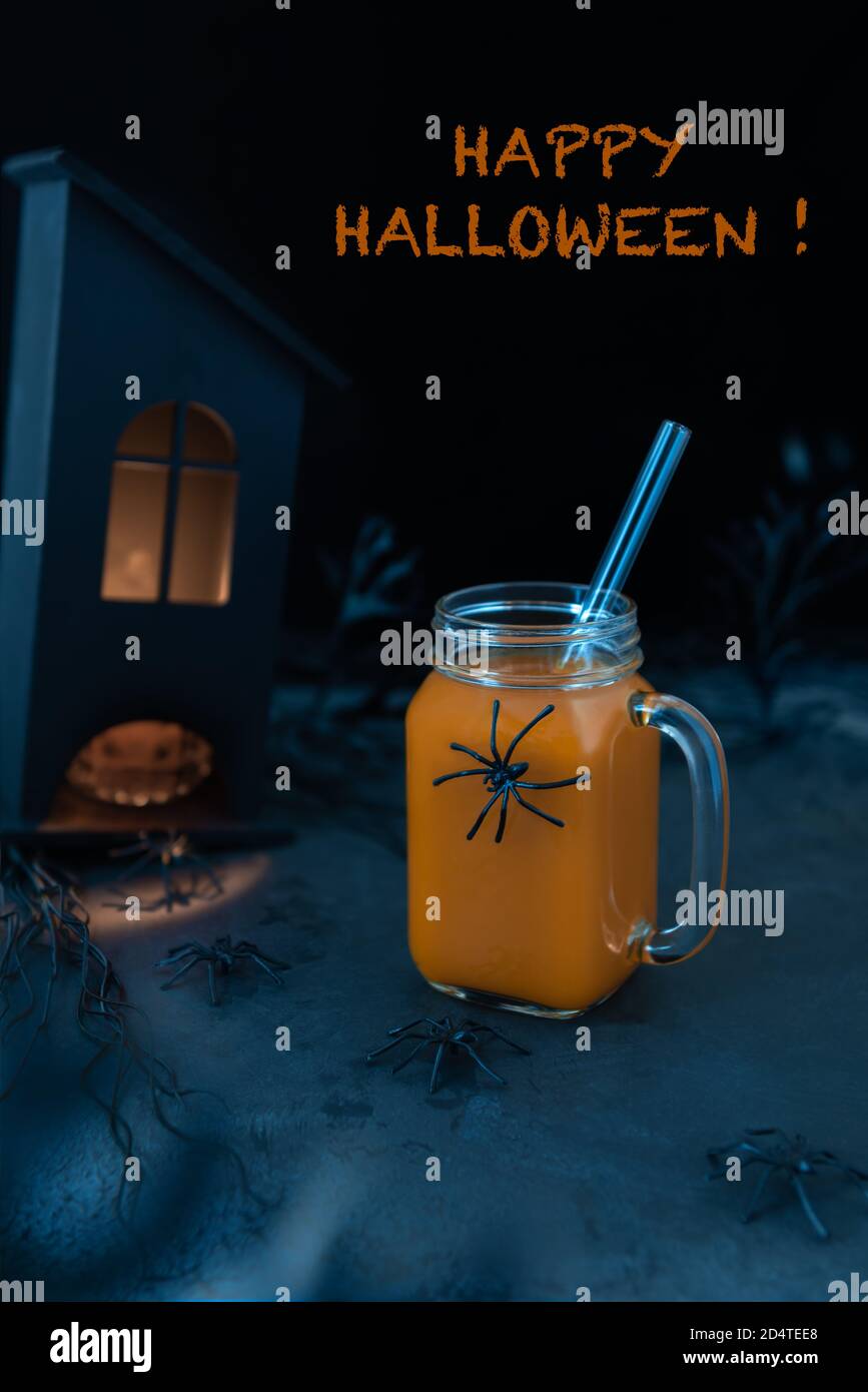 Halloween scene with pumpkin mocktail, spiders, spooky black house with light in windows and plants. Happy Halloween orange greeting text. Scary silho Stock Photo