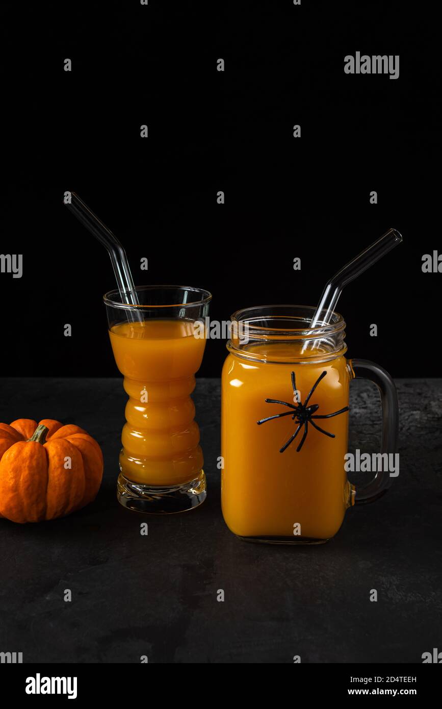 Non-alcoholic pumpkin mocktail in drinking glass and square jar with glass straws and whole orange pumpkin on black textured surface. Halloween spider Stock Photo