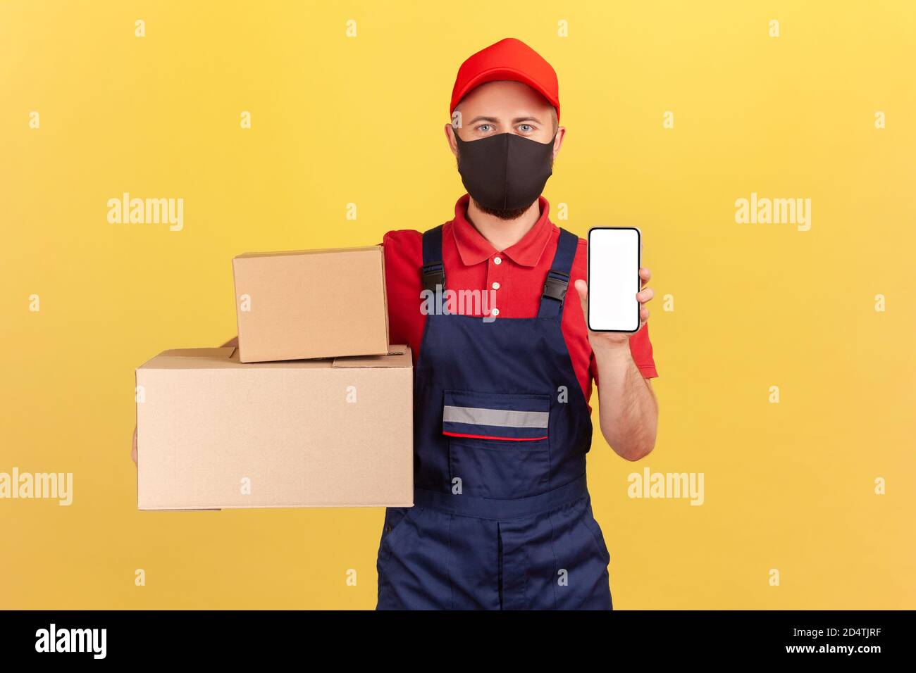 Courier in uniform and protective mask on face holding showing cardboard boxes and smartphone with empty screen, application for delivery, advertising Stock Photo