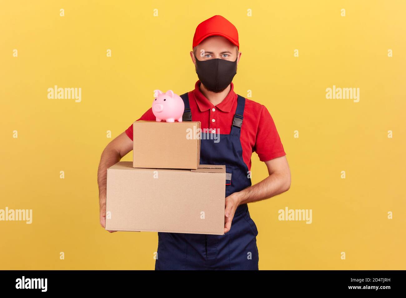 Delivery man in uniform and protective mask holding cardboard parcels with piggy bank on top, save money ordering delivery, advertising. Indoor studio Stock Photo