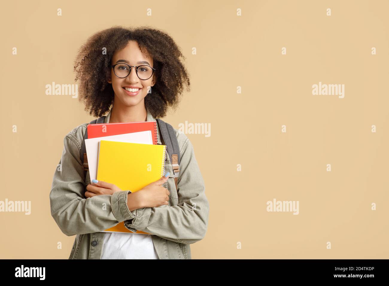 Preparation for lessons. Studio shot of glad african american female student in glasses with notepads, looks at camera, isolated on light background Stock Photo