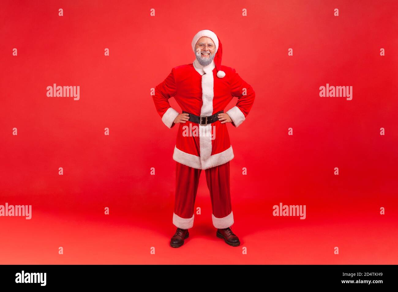 Full length portrait of positive happy aged man in santa claus costume standing holding hands akimbo, preparing for winter holidays and toothy smile. Stock Photo