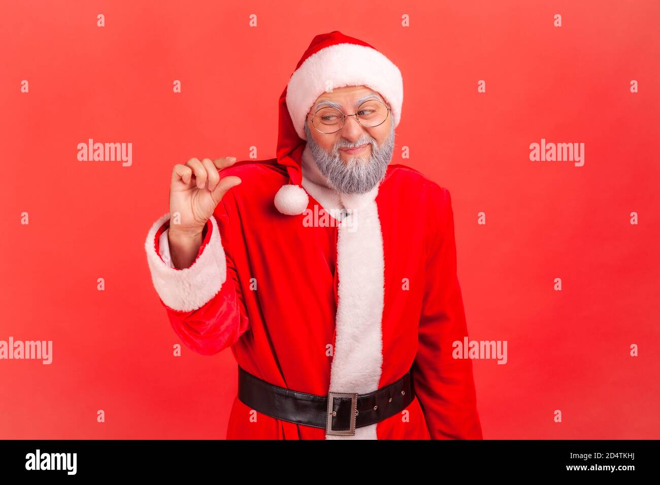 Portrait of dissatisfied elderly gray bearded man in santa claus costume showing a little gesture with fingers, skeptical about winter holidays magic. Stock Photo