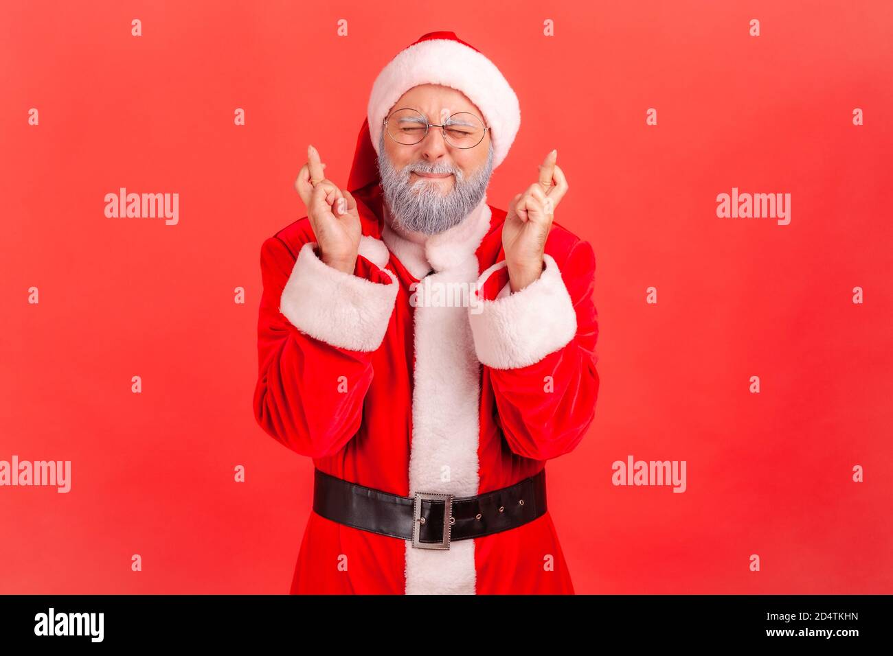 Portrait of dreamy elderly man in santa claus costume closing eyes and crossing fingers making a wish, christmas magic, winter holidays. Indoor studio Stock Photo