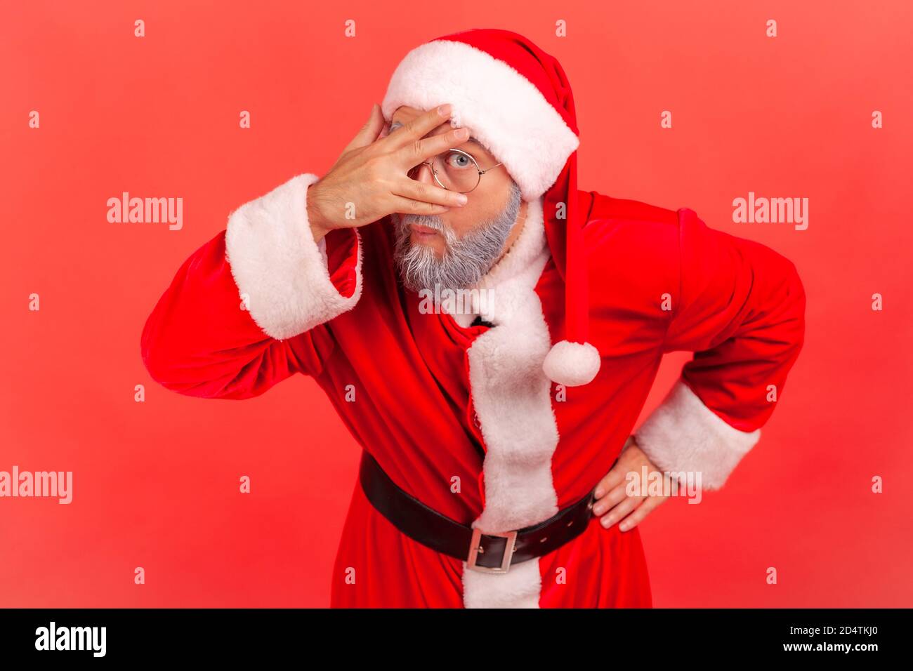 Portrait of curious nosy man in santa claus costume pretending to close eyes with hand attentively peeping through fingers, spying. Indoor studio shot Stock Photo