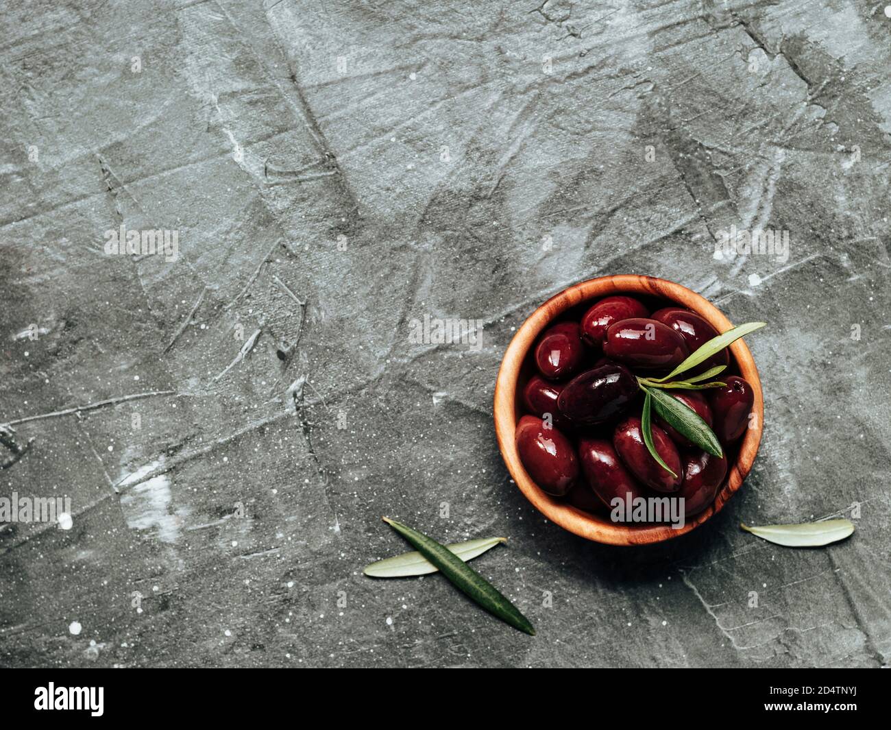 Kalamata olives on gray textured background. Red kalamata olives in olive wooden bowl with fresh green olives leaves, top view or flat lay. Copy space for text or design. Stock Photo