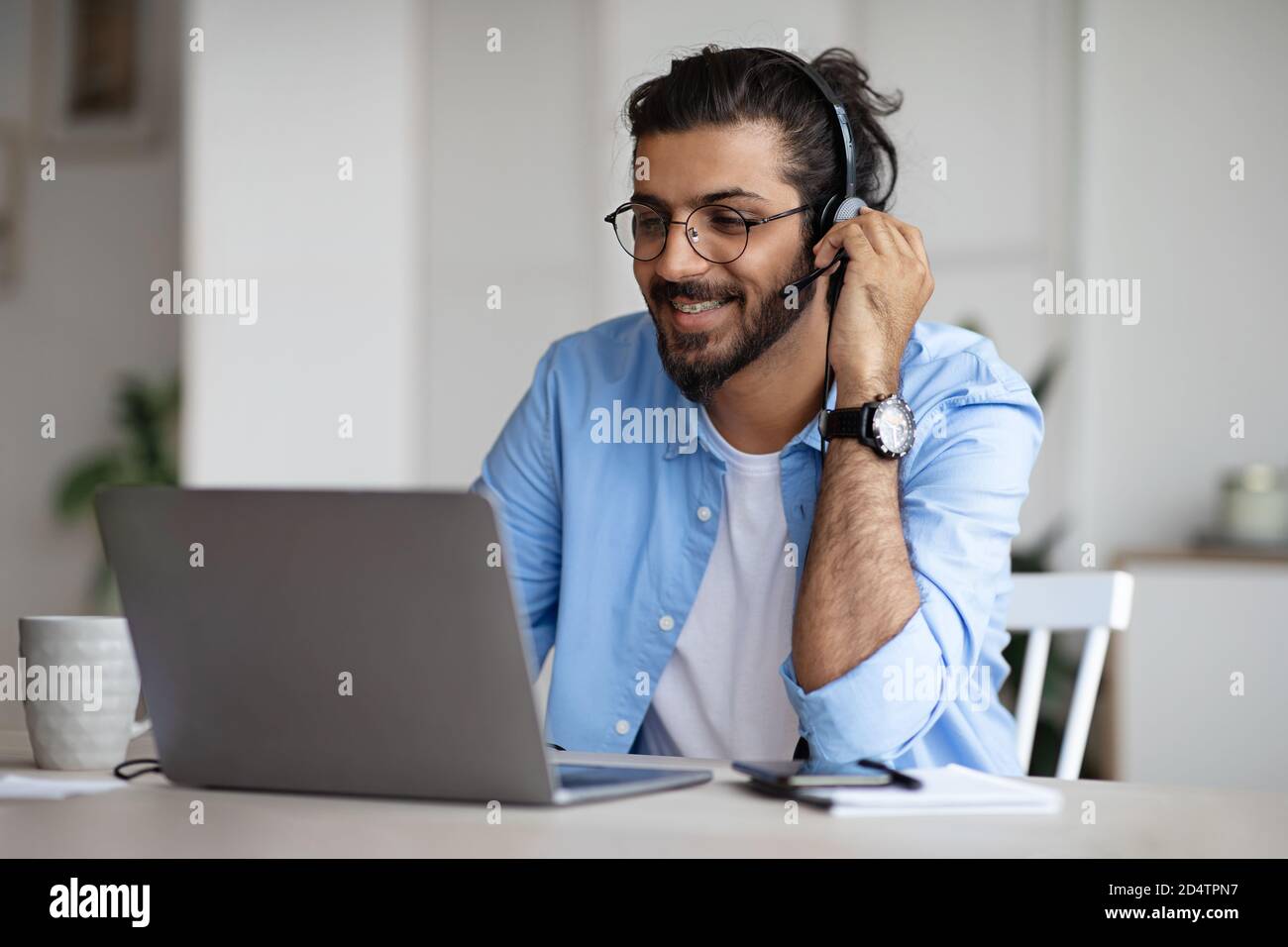 Smiling Young Indian Guy Watching Webinar On Laptop, Wearing Headset And Eyeglasses Stock Photo