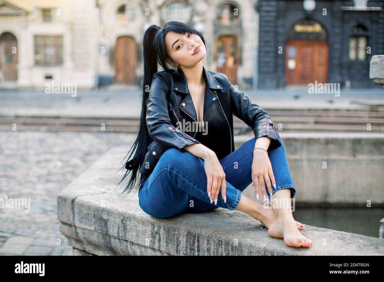 Beautiful young brunette Asian woman, dressed in jeans and black leather  jacket, posing in the center of the old European city, sitting on the stone  Stock Photo - Alamy