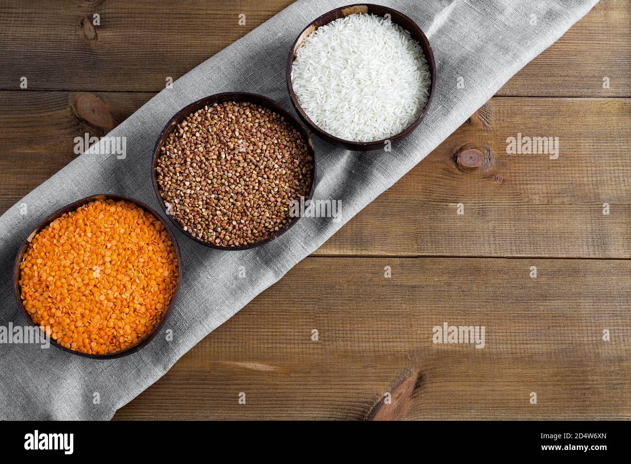 Three full bowls of raw cereals buckwheat, rice, lentils on a canvas napkin on a wooden background flat lay with place for text. Different types organ Stock Photo
