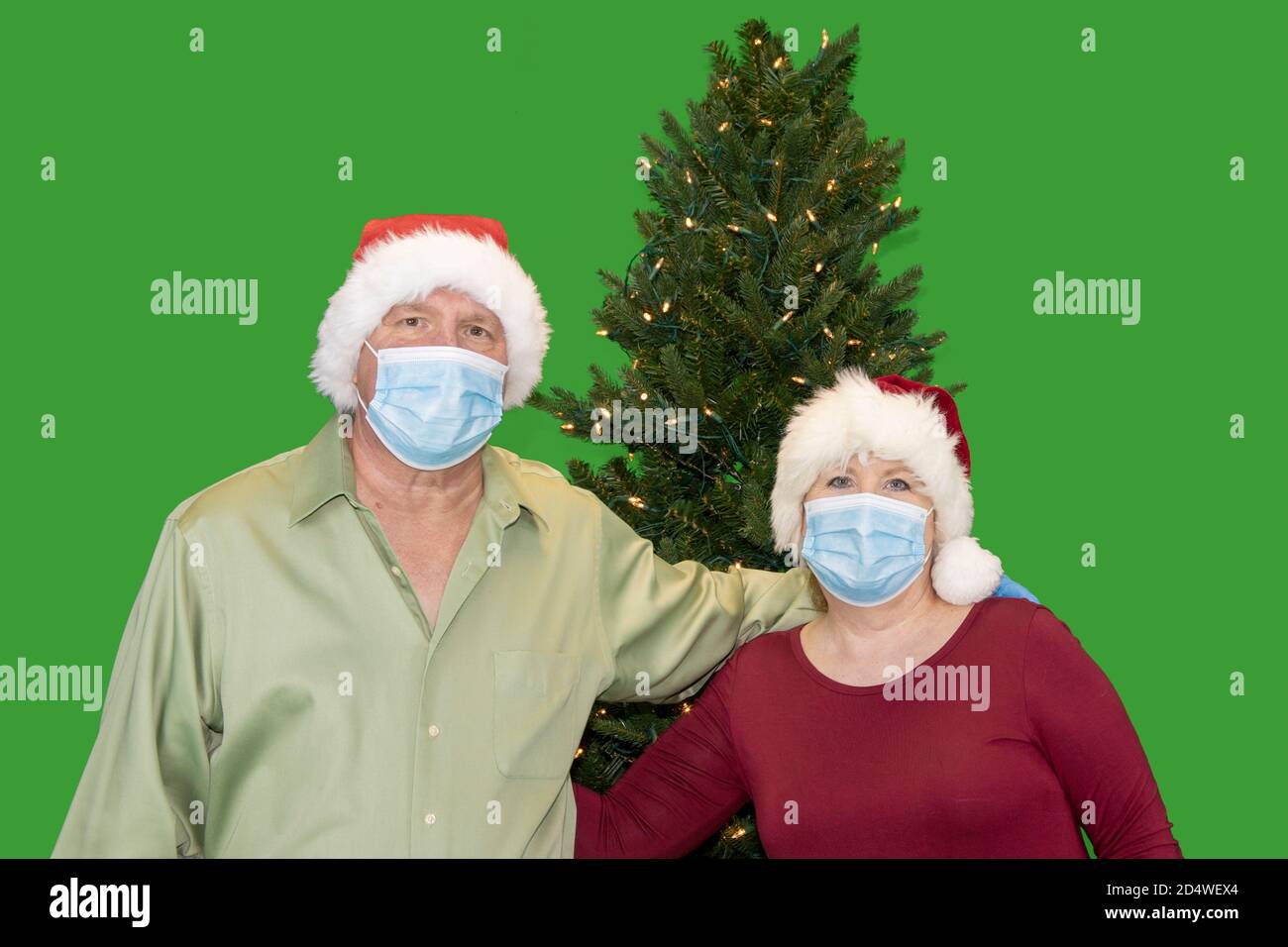 Older couple dressed in stocking hats wearing masks posing in front of Christmas tree with copy space on green screen. Stock Photo