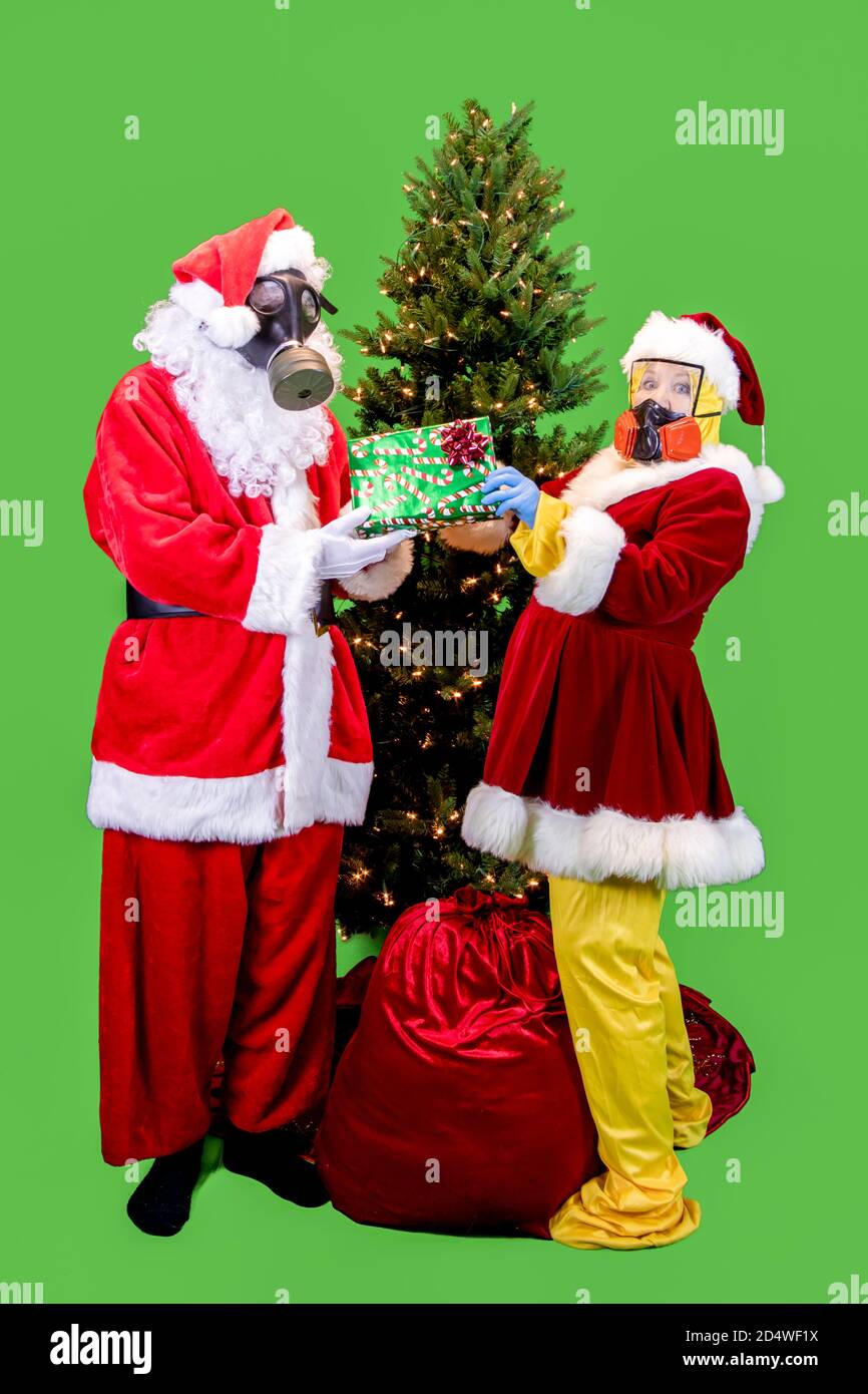 Santa Claus and the Mrs. dressed in ppe, or personal protective gear, including gloves, masks and suits exchanging gifts under Christmas tree with rem Stock Photo