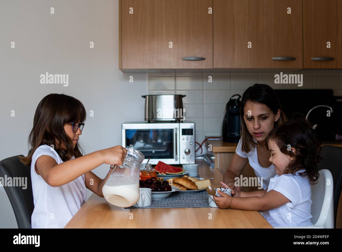 Caring young Caucasian mother having breakfast at the kitchen table with her two cute daughters Stock Photo
