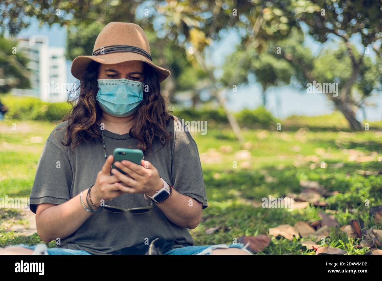 Portrait of a woman with a hat looking at her mobile that is protected with a medical mask and is outdoors. New normal. Copy space Stock Photo
