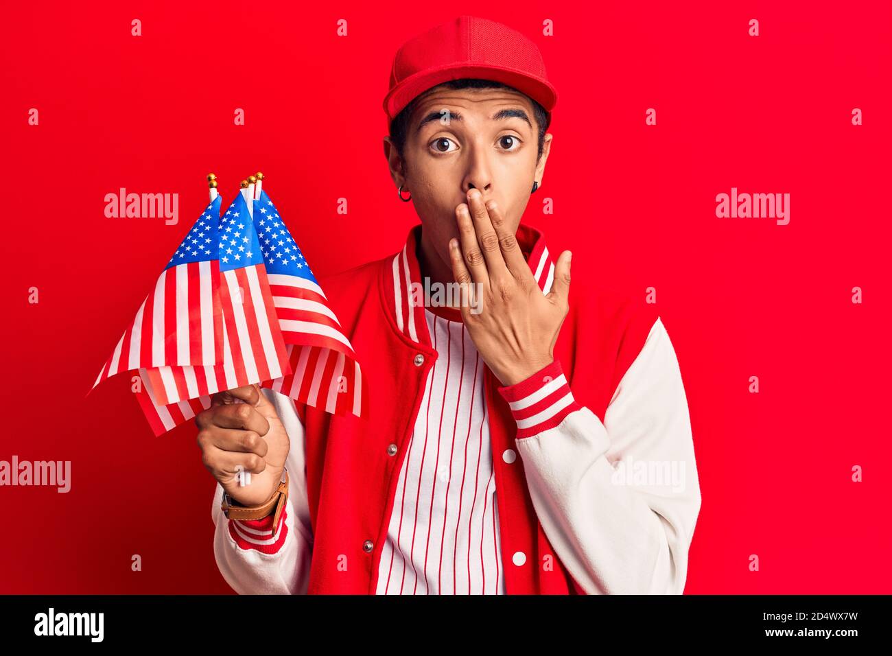 Young african amercian man wearing baseball uniform holding america flags covering mouth with hand, shocked and afraid for mistake. surprised expressi Stock Photo