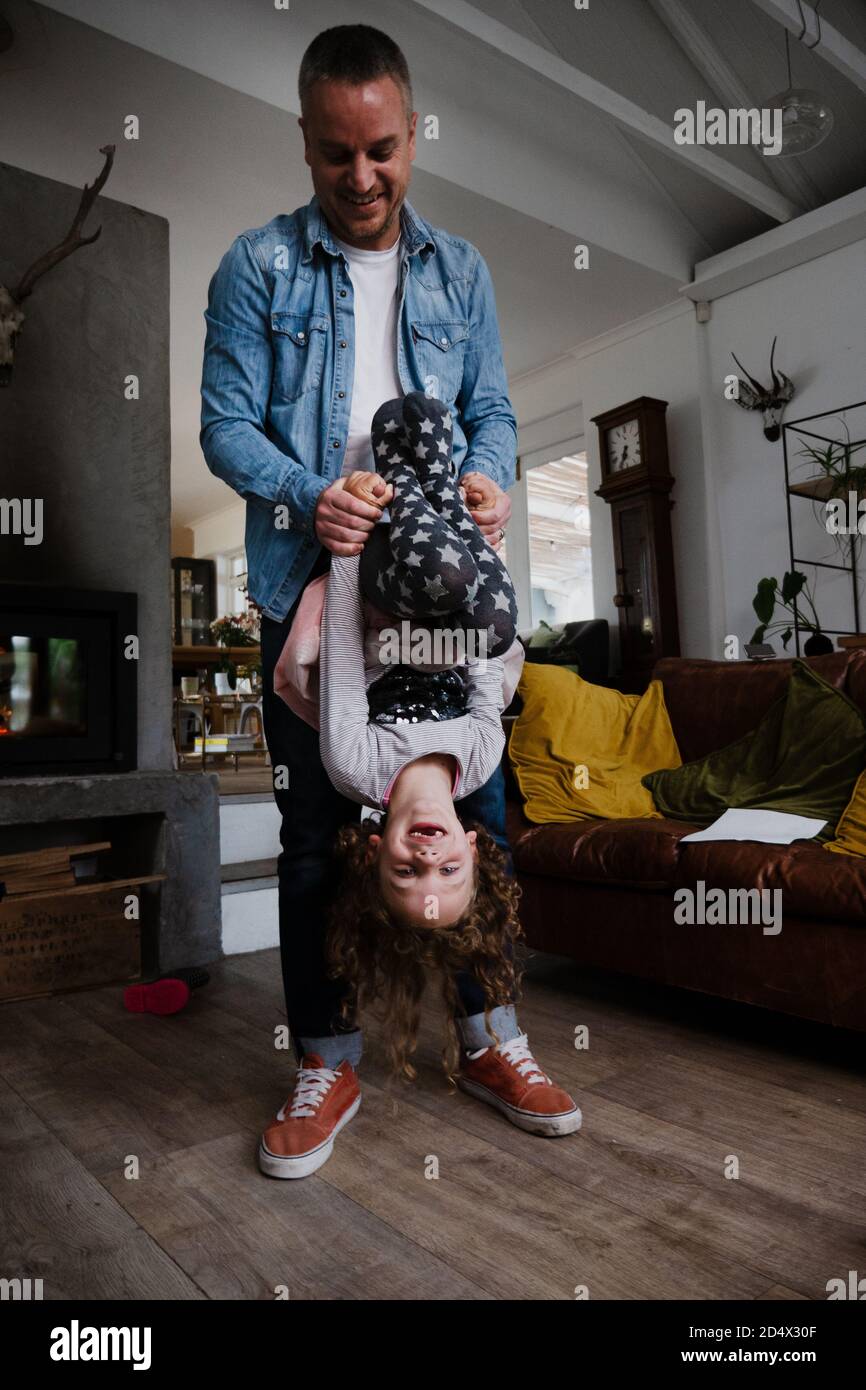 Dad and daughter bonding in living room hanging upside down with wide smile. Stock Photo