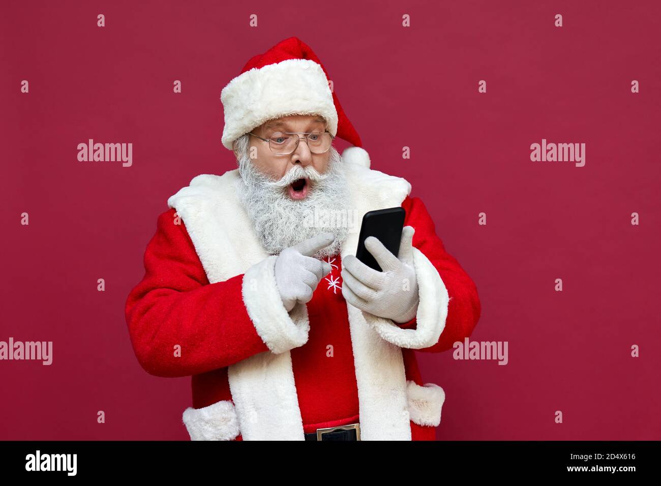 Surprised Santa Claus using smart phone isolated on red background. Stock Photo