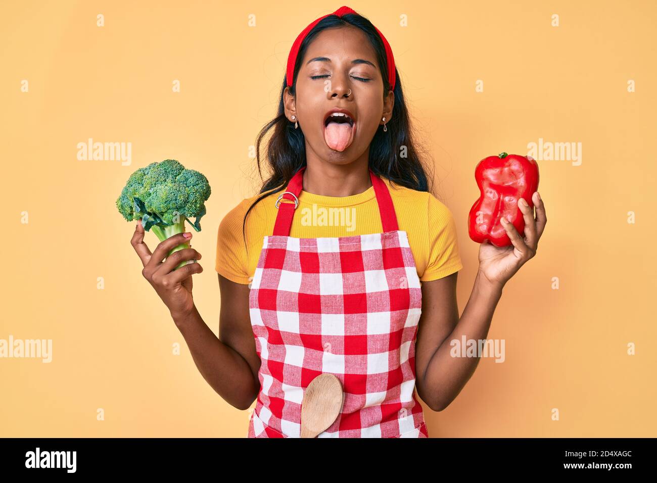 Young indian girl wearing apron holding broccoli and red pepper sticking tongue out happy with funny expression. Stock Photo